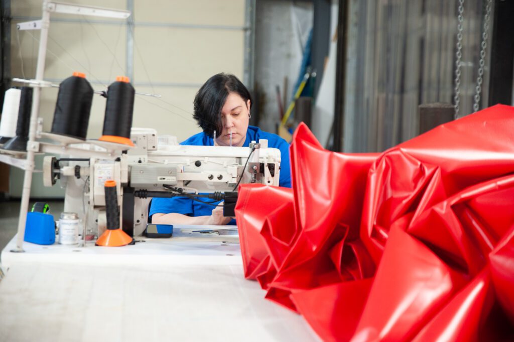A woman with short black hair operates a sewing machine, hemming a red tarp