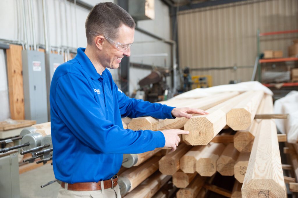 A man in safety goggles and a blue shirt stands next to a pile of wooden poles, pointing to the end grain